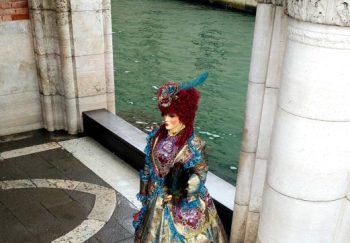 A person in an elaborate Venetian costume stands by historic architecture beside a canal in Venice.