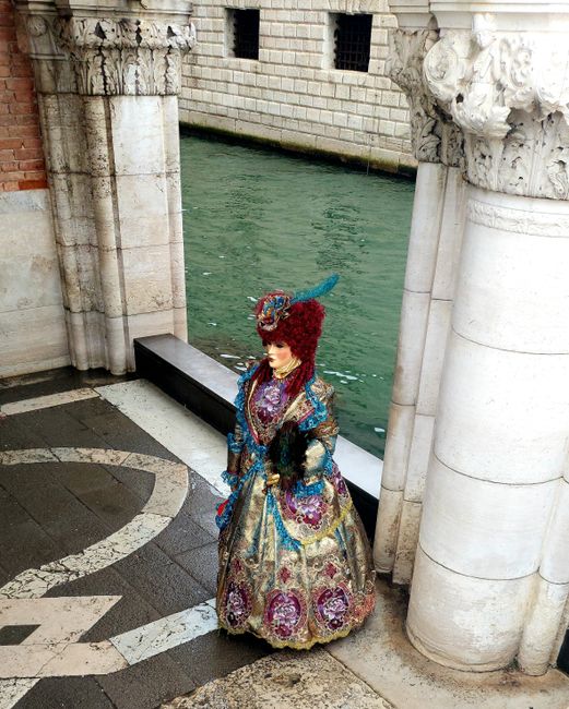 A person in an elaborate Venetian costume stands by historic architecture beside a canal in Venice.