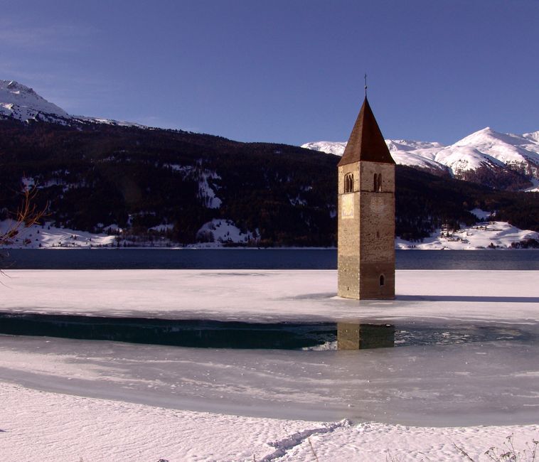 Winter view of the submerged bell tower in Lake Reschen, Italy, amidst snow-covered mountains.