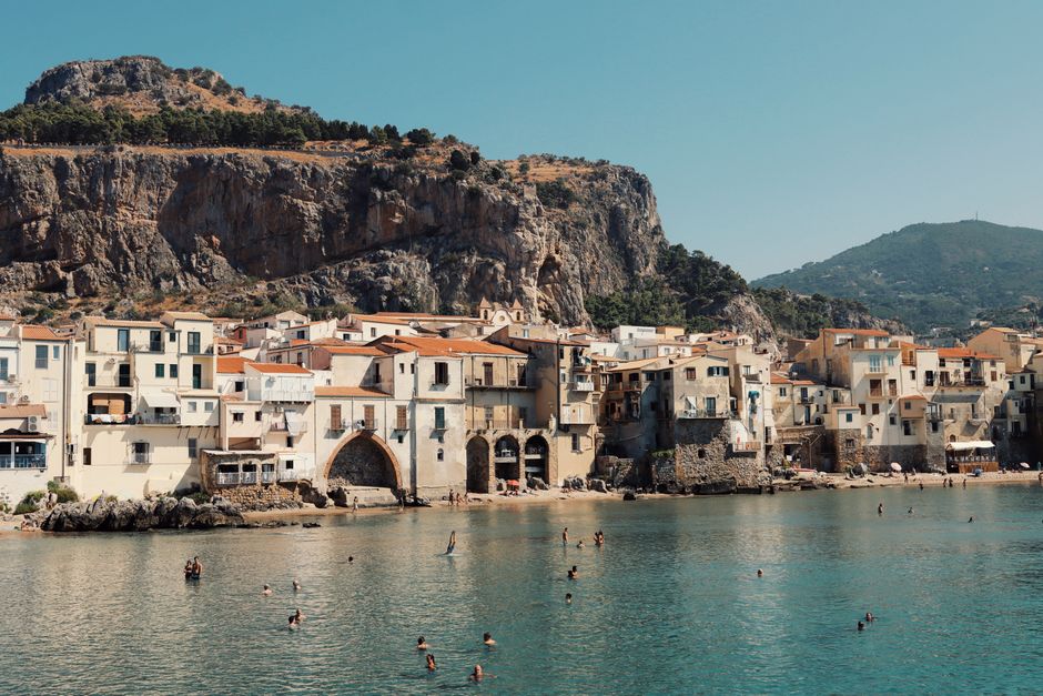 Swimmers enjoy the water with Cefalù's historic seaside buildings and rocky cliffs in Sicily, Italy.