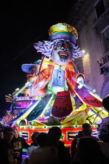 Colorful lit carnival float featuring a clown, drawing crowds at night.