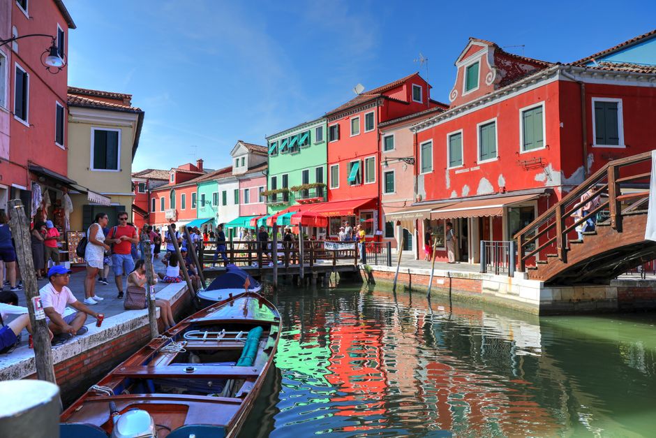 Colorful houses line the canal in Burano, Italy, with tourists enjoying a sunny day.