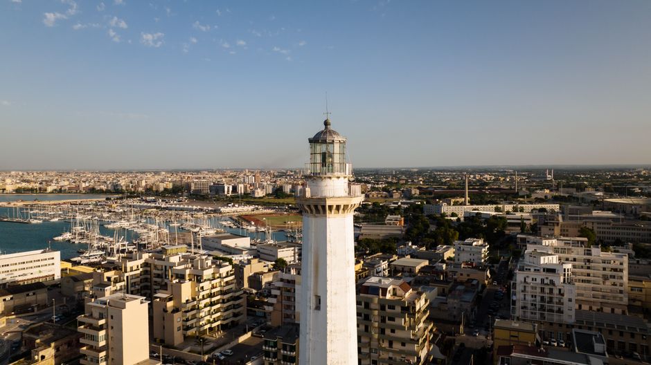 A stunning drone shot of a lighthouse in an urban coastal cityscape under a clear blue sky.