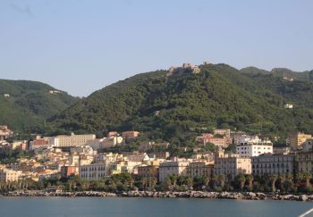 Panoramic view of Salerno's coastal cityscape flanked by lush green hills under a clear sky.