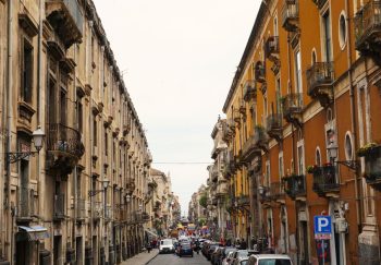 Charming city street with historic buildings and ornate balconies, bustling with vehicles and pedestrians.