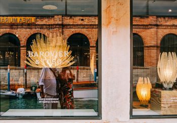 Photo of a store window showcasing unique light designs from Barovier & Toso, reflecting the vibrant cityscape.