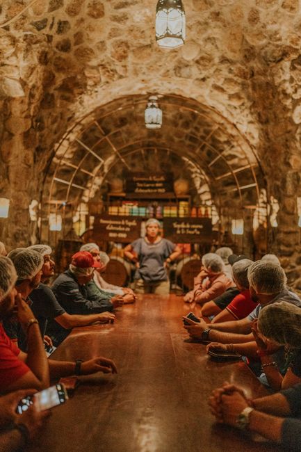 Group of tourists at a wine tasting event in a stone cellar, led by a guide.