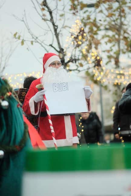 Santa Claus holding a sign outdoors with lights and people in a festive atmosphere.