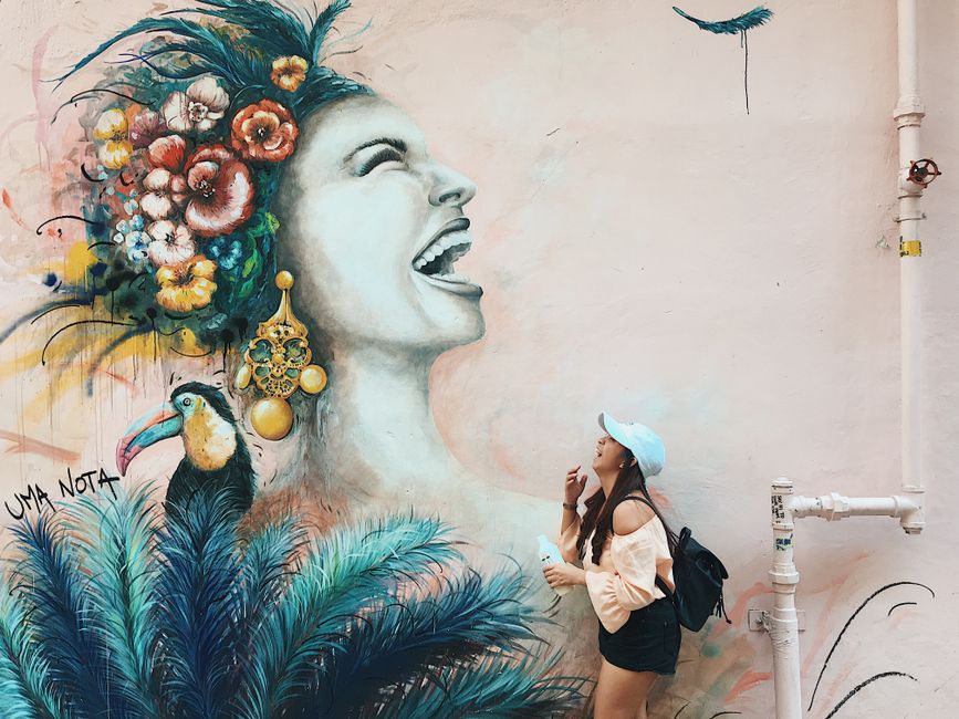 A young woman poses beside a colorful mural featuring a floral headdress and a toucan.