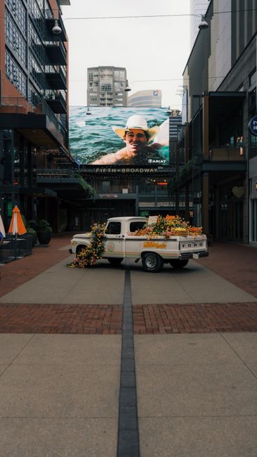 A vintage truck filled with flowers parked in an urban street setting with a large billboard overhead.