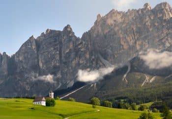 A serene view of a church against the majestic Dolomites mountains with lush greenery and clouds.