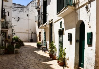 Sunlit alleyway in a classic Puglian town with whitewashed buildings and plants.