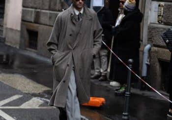 A man in a trench coat walks down a rainy street in Milan, showcasing urban fashion.