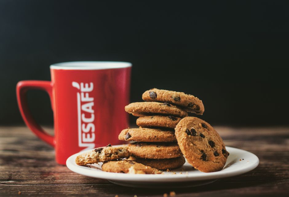 Stack of chocolate chip cookies on a plate with a red coffee cup on a dark background.