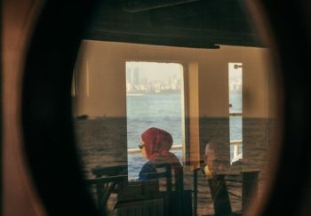 Two passengers viewed through a window reflection on a ferry crossing a tranquil sea.