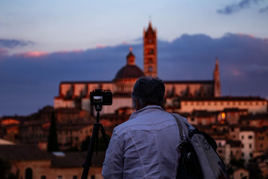 A photographer sets up a camera to capture the scenic skyline of Siena at dusk, with a cathedral in the background.
