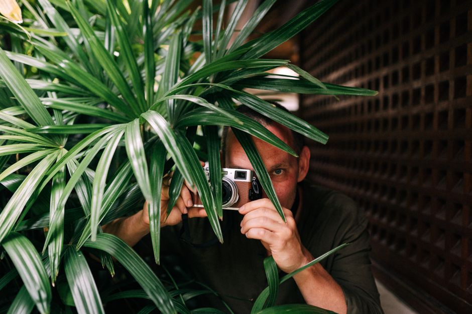 A man hides behind green foliage holding a camera, creating a playful and mysterious scene.