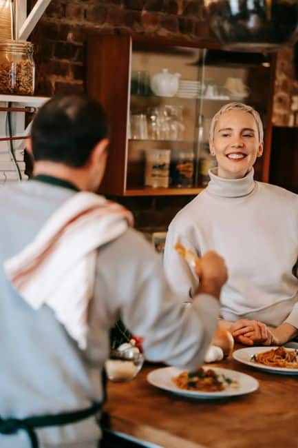 Happy adult female in casual clothes smiling while eating delicious spaghetti with tomato sauce with anonymous husband at home
