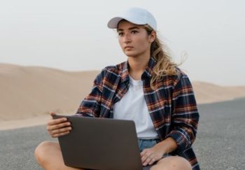 Young woman using laptop on a desert road, wearing casual attire and hat, symbolizing remote work.