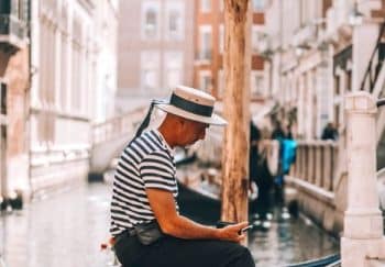 A gondolier in Venice relaxes on a bench by the canal, wearing a striped shirt and boater hat.