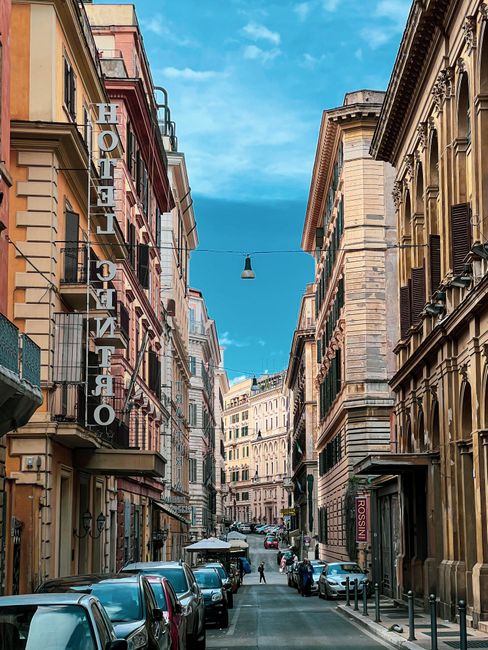 A picturesque Roman street lined with historic buildings and parked cars under a clear sky.