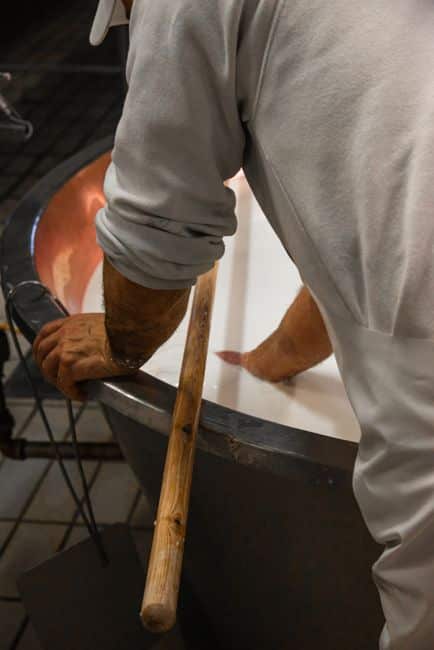 A man works with dairy ingredients in a large industrial vat, stirring with a wooden pole.