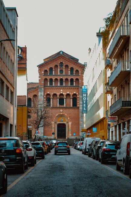 Street view of a historic church facade in Milan, Italy with parked cars lining both sides.