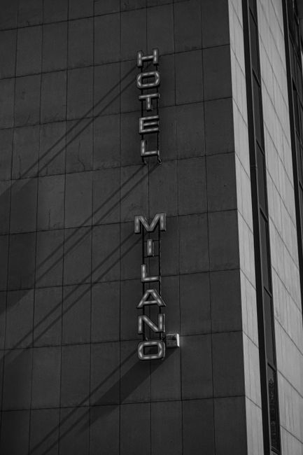 Monochrome image of Hotel Milano exterior with bold vertical signage and dramatic shadows.