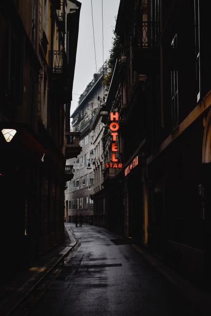 Dark, narrow street in Milan featuring a glowing neon hotel sign, creating a moody urban atmosphere.