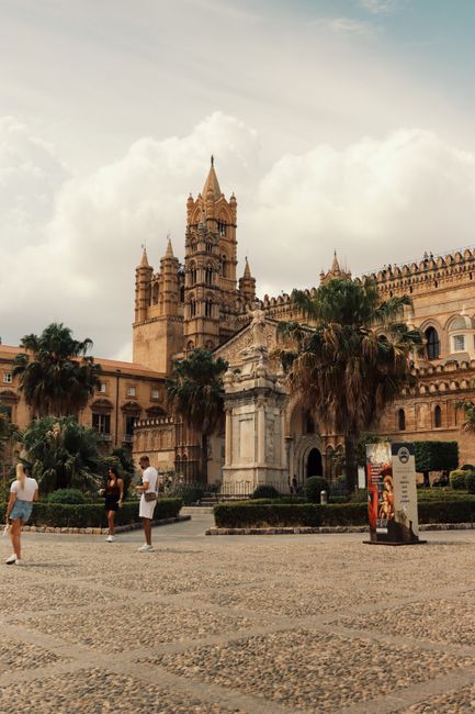 Beautiful view of Palermo Cathedral and silhouetted people, with palm trees and clouds in Sicily.