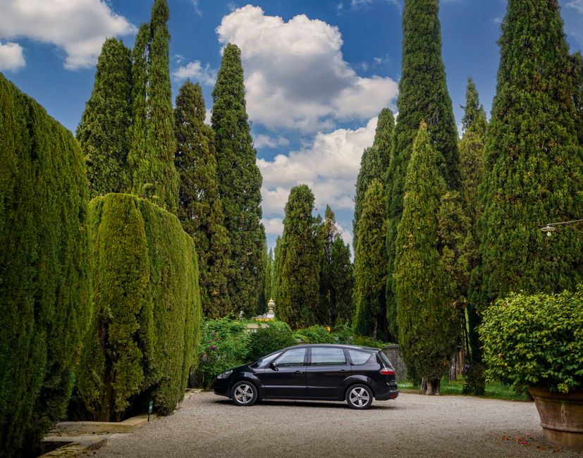 A black car parked in a beautiful Tuscan garden surrounded by lush cypress trees.