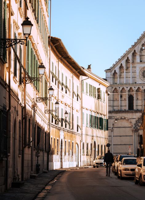 A man walks down a sunlit street in Pisa, Tuscany with historic buildings and a clear blue sky.