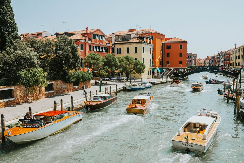 Colorful motorboats navigate a bustling Venetian canal lined with vibrant historic buildings and lush greenery.