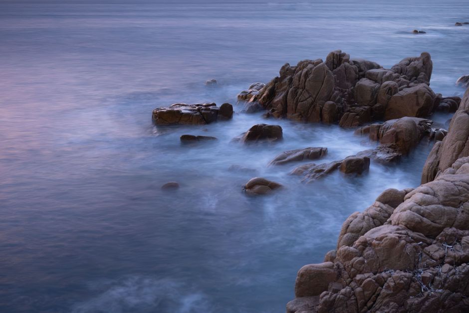 Serene early morning view of rocky coastline in Sardinia with calm waves and pastel sky.