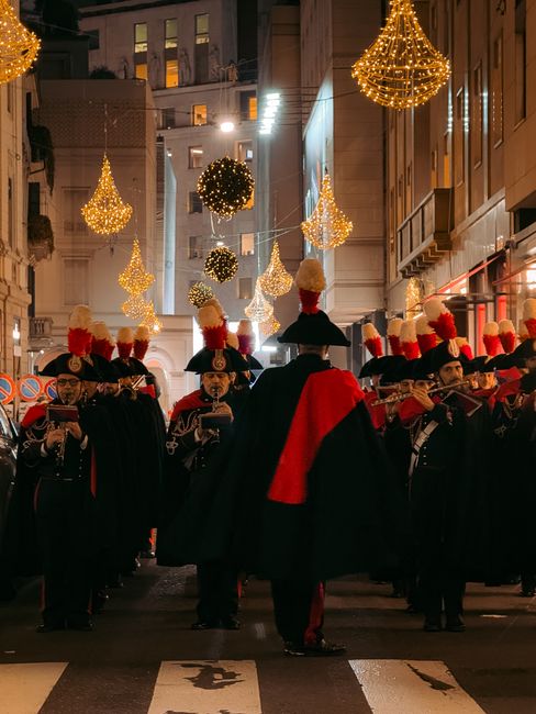 Nighttime parade in Milan featuring a marching band in festive uniforms under glowing street decorations.