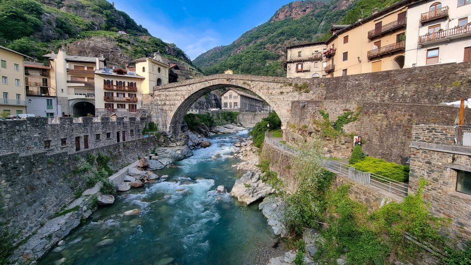 Historic stone bridge crossing a gentle river in scenic Bard, Italy, surrounded by picturesque buildings and lush greenery.