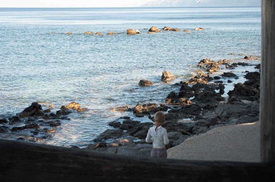 A young child stands by the rocky shores of Cala Gonone, Italy, gazing at the tranquil sea.