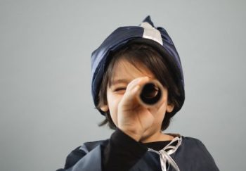 Child in costume of astronomer with stars squinting while looking at camera through toy telescope on gray background