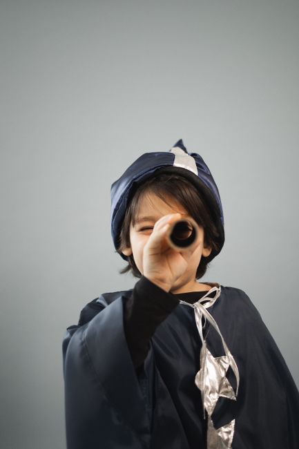 Child in costume of astronomer with stars squinting while looking at camera through toy telescope on gray background
