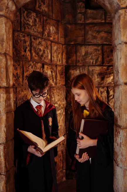 Two children dressed as wizards reading books in a stone corridor.