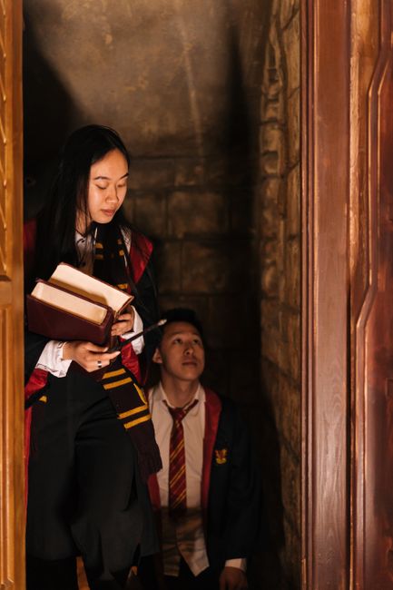 Two young adults in wizard attire step through a dimly lit stone doorway.
