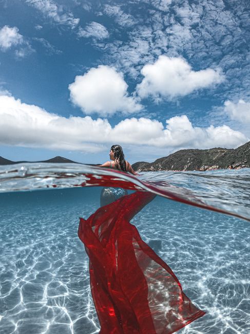 A woman with a flowing red fabric swimming in crystal clear water under a bright blue sky.