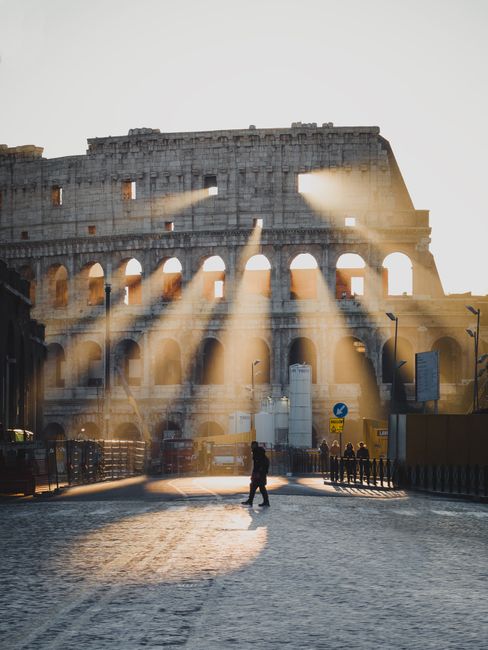 A person walking in front of the Colosseum in Rome, Italy, with sunrays filtering through the arches at sunrise.